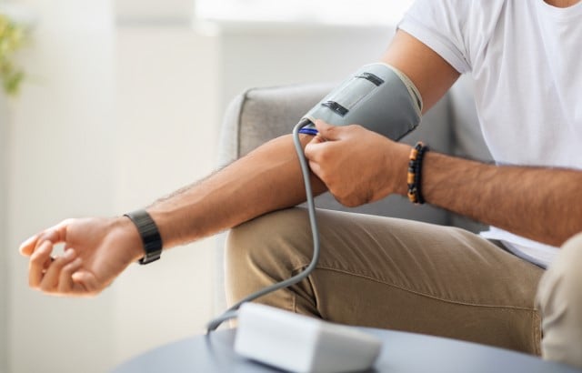 A white man's arm is outstretched with a grey blood pressure cuff around the upper part of the arm. He has a black watch on his wrist, tan trousers and a white shirt, but you can't see his face